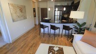 This single-floor space showcases a thoughtfully designed open-concept layout that seamlessly connects the kitchen and living areas. Rich hardwood flooring flows throughout, lending warmth and continuity to the space. The kitchen is a standout feature, appointed with dark espresso cabinetry that extends to the upper walls, providing ample storage. Stainless steel appliances, including a full-size refrigerator, add a sleek, modern touch, while a classic tile backsplash introduces subtle texture and depth. The centerpiece of the kitchen is a curved breakfast bar topped with a bold dark granite countertop, accommodating three contemporary bar stools with clean lines and dark upholstery. A decorative bowl rests atop the bar, adding an artistic accent. Pendant lighting hangs above, casting a warm, focused glow over the workspace and dining area. The living area benefits from the open sightlines, creating a spacious and airy feel. A large wall-mounted artwork and a cozy area rug with a cowhide accent piece ground the living space with character. The overall ambiance is modern and inviting, balancing functional design with stylish, cohesive finishes throughout.