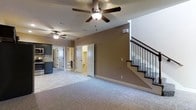 This two-story home offers a functional and comfortable layout across both levels. The main floor centers on a spacious carpeted living area finished with warm taupe-toned walls and white trim, creating a neutral and inviting atmosphere. Ceiling fans are installed throughout, promoting airflow and comfort. A staircase with dark wood treads and decorative metal balusters with diamond detailing provides access to the upper level, serving as a subtle architectural accent within the space.

Adjacent to the living area, the kitchen features dark cabinetry with a mosaic tile backsplash and is equipped with appliances, offering a practical cooking space. The main level also includes at least one bathroom, visible through open doorways, finished with white wainscoting and light-toned flooring. Multiple interior doors with paneled detailing maintain a consistent design language throughout the home. The open sightlines between the living area, kitchen, and adjoining rooms contribute to a connected, easy-to-navigate floor plan suited for everyday living.