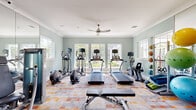 This single-story fitness facility offers a well-appointed workout space across a generously sized floor plan, designed to accommodate a comprehensive range of exercise activities. The interior is finished with soft sage-green walls, white crown molding, and a vibrant multicolored geometric-patterned carpet that adds energy to the environment. Ceiling fans are positioned throughout, promoting air circulation and comfort during workouts.

Natural light flows abundantly through multiple large windows lining the walls, offering views of lush outdoor greenery and creating an open, airy atmosphere. Full-length mirror panels are installed along one wall, visually expanding the space and providing functional feedback during strength training.

The cardio zone is well stocked with multiple treadmills, elliptical trainers, stationary bikes, and a rowing machine, all equipped with individual display screens. A dedicated strength training area features a multi-station cable machine, a weight bench, and a tiered dumbbell rack offering a range of free weights. Functional training accessories include stability balls, medicine balls, resistance bands, and yoga mats stored on organized racks. Recessed lighting supplements natural light, ensuring the space remains bright and practical throughout the day.
