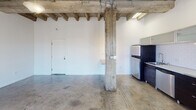 Located on Beverly Boulevard in Los Angeles, this single-floor space showcases a compelling industrial aesthetic that blends raw architectural elements with modern finishes. The open main area is defined by striking exposed wood beam ceilings supported by original structural columns, lending the space a loft-like character with generous vertical volume. Polished and raw concrete floors run throughout, adding to the industrial ambiance while providing a durable, easy-to-maintain surface.

The kitchen is well-appointed and runs along one wall, featuring dark cabinetry contrasted by a crisp white subway tile backsplash and light countertops. Upper cabinets with frosted glass-front doors add a contemporary touch, while stainless steel appliances — including a gas range, dishwasher, and refrigerator — complete the functional layout.

The remaining rooms offer bright, white-walled interiors with concrete floors and windows fitted with horizontal blinds that allow natural light to filter in. A separate room with a smoother ceiling finish provides a quieter, more enclosed feel compared to the open main area. The bathroom is clean and straightforward, equipped with a pedestal sink, toilet, and wall mirror. Overall, the space presents a versatile industrial character well-suited for a variety of uses.