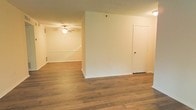 This single-floor residence offers a clean, open layout designed for comfortable everyday living. The interior is finished with warm-toned hardwood-style flooring that flows seamlessly throughout the main living areas, adding a sense of continuity and warmth to the space. Crisp white walls and neutral tones create a bright, airy atmosphere that serves as a versatile backdrop for a variety of interior styles. The main living area is spacious and well-proportioned, with a partial wall division that subtly separates functional zones while maintaining an open feel. A ceiling fan overhead provides both lighting and airflow, enhancing comfort in the connected room. A interior door off to one side provides access to an additional room or utility space, contributing to the home's overall practical layout. Recessed floor vents suggest a central HVAC system for year-round climate control. The understated, move-in-ready design of this single-story home makes it an ideal canvas for personalization, offering a solid foundation of quality finishes and a thoughtfully arranged floor plan.
