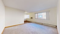 This single-floor residence offers a well-organized layout with a variety of functional spaces throughout. The generously sized living area is finished with soft wall-to-wall carpeting and features multiple windows that allow natural light to fill the room, with views of the surrounding brick building exterior. A baseboard heating system and a wall-mounted air conditioning unit provide year-round climate control. Warm wood-toned baseboards and trim add a touch of character throughout the space.

The galley-style kitchen is efficiently designed with white cabinetry lining one wall, complemented by laminate countertops, a single-basin sink, and white appliances including a full-size refrigerator. Rich wood-look flooring runs the length of the kitchen, adding warmth to the space.

The bedroom features carpeted flooring, a painted cinder block accent wall that gives the room a sturdy, grounded feel, and a sliding-door closet fitted with built-in shelving and a hanging rod for ample storage. An additional open shelving unit is also present nearby.

The bathroom is finished with classic mosaic tile flooring in a neutral palette, a full soaking tub, a toilet, and a window that brings in natural light. A warm wood door frames the entrance to this bright and functional space.
