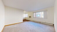 This single-floor residence offers a well-organized layout with a variety of functional spaces throughout. The generously sized living area is finished with soft wall-to-wall carpeting and features multiple windows that allow natural light to fill the room, with views of the surrounding brick building exterior. A baseboard heating system and a wall-mounted air conditioning unit provide year-round climate control. Warm wood-toned baseboards and trim add a touch of character throughout the space.

The galley-style kitchen is efficiently designed with white cabinetry lining one wall, complemented by laminate countertops, a single-basin sink, and white appliances including a full-size refrigerator. Rich wood-look flooring runs the length of the kitchen, adding warmth to the space.

The bedroom features carpeted flooring, a painted cinder block accent wall that gives the room a sturdy, grounded feel, and a sliding-door closet fitted with built-in shelving and a hanging rod for ample storage. An additional open shelving unit is also present nearby.

The bathroom is finished with classic mosaic tile flooring in a neutral palette, a full soaking tub, a toilet, and a window that brings in natural light. A warm wood door frames the entrance to this bright and functional space.