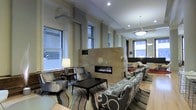 This single-floor space showcases a thoughtfully designed interior that blends contemporary elegance with comfortable functionality. The centerpiece of the main lounge area is a sleek, linear double-sided fireplace encased in a tall stone surround, serving as a dramatic architectural divider between two distinct seating zones. On one side, plush gray armchairs are arranged around a dark coffee table atop a patterned area rug, accompanied by a classic table lamp that adds warm ambient lighting. The opposite side features bold patterned sofas and additional seating, creating an inviting social area. Tall windows on multiple walls flood the interior with generous natural light, offering views of the surrounding urban environment. Recessed ceiling lighting and pendant fixtures — including a notable bowl-shaped pendant and a geometric hanging light — provide layered illumination throughout the space. The flooring transitions between hardwood and area rugs, adding texture and warmth. Neutral wall tones, refined millwork detailing, and carefully curated furnishings contribute to a polished, upscale ambiance suited for both relaxation and social engagement.