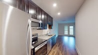 This well-appointed single-floor residence offers a thoughtfully designed layout with modern finishes throughout. The kitchen serves as a central highlight, featuring rich dark espresso cabinetry extending to the ceiling for ample storage, granite countertops, a stainless steel sink, and a full suite of stainless steel appliances including a refrigerator, electric range, built-in microwave, and dishwasher. Recessed lighting illuminates the space, which flows seamlessly into the open living area via warm hardwood-style flooring. The living area benefits from natural light streaming through a glass-paned door that leads to an outdoor balcony or patio, enhancing the sense of openness. The bedroom is finished with plush neutral carpeting and features a window with views of surrounding greenery, creating a calm and comfortable retreat. The bathroom is outfitted with a dark vanity cabinet, a white countertop with a modern brushed-nickel faucet, a large mirror, and distinctive black-and-white mosaic tile flooring that adds a classic design touch. A dedicated in-unit laundry area houses a stacked washer and dryer, providing everyday convenience. Throughout the residence, clean white walls and consistent flooring tie each space together cohesively.