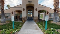 The Newport Village Apartments clubhouse is a single-story community facility designed to serve residents with a range of amenities. The exterior presents a stucco facade with stone accents framing a grand arched entryway, complemented by manicured flower beds, palm trees, and a well-maintained walkway that creates an inviting first impression.

Inside, the fitness center stands out as a highlight of the facility. The room features soaring vaulted ceilings with a ceiling fan overhead, fostering an open and airy atmosphere. Three arched windows along one wall flood the space with natural light and offer views of the surrounding greenery. Light blue walls are accented with bold geometric yellow stripe designs, adding a modern, energetic feel. Durable hardwood-style flooring supports strength training equipment and benches positioned throughout the room.

Adjacent to the fitness area, the lounge provides a comfortable retreat with warm neutral-toned walls accented by white chair rail molding. Light hardwood floors, recessed lighting, and a ceiling fan contribute to a relaxed ambiance. A plush sectional sofa, area rugs, and a modular shelving unit furnish the space, making it ideal for residents to unwind or socialize.