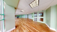 This well-appointed fitness and yoga studio offers a welcoming, open-plan layout designed to accommodate a variety of wellness activities. The space is immediately distinguished by its warm, honey-toned hardwood flooring that extends throughout the entire area, providing both a visually appealing and practical surface for exercise and movement.

The walls are finished in a calming sage green, contributing to a serene and energizing atmosphere. A striking wall of floor-to-ceiling windows runs along one side of the studio, flooding the interior with abundant natural light and offering expansive panoramic views of the surrounding area. Sheer white curtains on grommet rods are installed along sections of the interior, offering flexible privacy or room division as needed.

Overhead, a drop-tile ceiling houses a series of recessed and flush-mounted panel light fixtures, ensuring consistent, bright illumination throughout the space. Climate control is provided via ceiling-mounted HVAC units integrated discreetly into the ceiling grid.

Glass-paneled interior doors with light green frames connect to adjacent areas, maintaining visual openness while delineating functional zones. Decorative touches, including yoga-themed wall decals, wall-mounted displays, and a sun ornament, reflect the studio's wellness-focused identity. Exercise equipment such as stability balls is also visible, underscoring the space's active, health-oriented purpose.