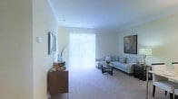 This two-floor residence offers a clean, understated aesthetic defined by neutral tones and classic architectural detailing. The living area is finished with wall-to-wall carpeting in a warm beige, contributing to a comfortable and quiet atmosphere. Crown moulding frames the ceiling, adding a refined touch to the otherwise minimalist design. A large sliding glass door fitted with vertical blinds serves as the room's focal point, drawing in abundant natural light and offering a visual connection to the outdoors. The walls are painted in a soft, pale green-white hue that keeps the space feeling open and airy. The living room accommodates a light grey sectional sofa paired with a glass-top coffee table, creating a practical and inviting seating arrangement. Decorative accents including framed artwork and a tall vase with branches add personality without overwhelming the space. A low wooden sideboard along one wall provides additional storage and display surface. The overall ambiance is calm and well-lit, with a straightforward layout that makes efficient use of the available space across both levels of the home.