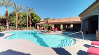 This outdoor amenity space showcases a resort-style pool area defined by its Mediterranean-inspired architectural design. The single-story clubhouse building features a distinctive terra cotta tile roof, warm stucco exterior walls, and arched architectural details that lend a cohesive, upscale aesthetic to the environment. The generously sized swimming pool has a freeform shape, giving it an organic, leisure-oriented character that invites relaxation. Adjacent to the main pool is a separate circular spa with stainless steel entry rails, offering a dedicated space for hydrotherapy. The surrounding pool deck is finished in a smooth concrete surface and is furnished with vibrant red chaise lounge chairs, providing ample space for sunbathing and outdoor lounging. A central palm tree planter island is positioned within the pool area, adding a tropical focal point and enhancing the resort atmosphere. Mature palm trees and lush greenery frame the perimeter, offering natural shade and privacy. Large multi-pane windows along the clubhouse facade allow natural light to enter the interior spaces while maintaining visual connection to the outdoor area. The entire setting is bathed in abundant natural sunlight beneath clear blue skies, reinforcing its warm-weather, leisure-focused character.
