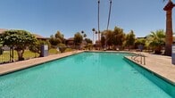 Located at 1950 North 43rd Avenue in Phoenix, Arizona, this outdoor amenity space showcases a generously sized swimming pool that serves as the centerpiece of the area. The pool features clear, turquoise water and is accessible via stainless steel entry rails, with a surrounding concrete deck that provides ample space for lounging and recreation.

A covered patio structure adjoins the pool area, offering a shaded retreat from the Arizona sun and furnished with lounge chairs for relaxed outdoor enjoyment. The entire pool area is enclosed by a wrought iron security fence, ensuring a safe and private environment for users.

Mature palm trees and lush greenery frame the space, providing a tropical aesthetic that complements the warm desert climate. Additional landscaping throughout the area includes manicured shrubs, grass lawns, and a variety of trees that create a refreshing contrast to the arid surroundings. Under the expansive, clear blue Phoenix sky, the outdoor setting exudes a bright, open ambiance year-round.

The surrounding residential buildings, featuring a mix of stucco and Spanish-tile architectural elements, are visible beyond the fencing, situating the pool area as a shared community amenity. This well-maintained outdoor space is thoughtfully designed to offer both relaxation and recreation in the heart of Phoenix.
