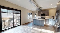 This modern single-floor space showcases a thoughtfully designed open-concept layout, where the living area and kitchen flow seamlessly together. The kitchen is a standout feature, equipped with sleek wood-grain cabinetry with matte black hardware, light quartz countertops, a central island, and a full suite of stainless steel appliances including a gas range, built-in microwave, dishwasher, and refrigerator. Stylish glass pendant lights hang above the island, while exposed industrial-style ductwork adds an urban character to the space. Light hardwood flooring runs throughout the main living and kitchen areas, complementing the neutral warm tones of the walls. Large black-framed windows and a sliding glass door allow generous natural light to fill the open living space, offering views of mature trees outside. The bedroom is generously sized with plush carpet, multiple windows, a ceiling fan with light fixture, and a built-in closet. Two bathrooms complete the unit: one full bath featuring a tub-shower combination with white hexagonal tile surround, wood-grain vanity, tall linen tower, large-format floor tile, and matte black fixtures, and a second bathroom with a clean, minimalist design featuring hexagonal mosaic tile flooring and matching black hardware.