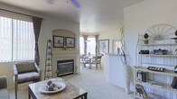 This single-floor residence offers a thoughtfully designed open-concept layout that seamlessly connects the living and dining areas, creating a spacious and inviting atmosphere. The living room serves as the heart of the home, anchored by a classically styled fireplace set within a tiled surround and framed by an arched alcove, above which a pair of coordinating framed artworks are displayed. Plush wall-to-wall carpeting in a warm neutral tone runs throughout, contributing to the overall sense of comfort and cohesion. A ceiling fan overhead adds both function and a subtle decorative touch. Decorative accents — including tall sculptural vases, a tiered display shelf, and natural branch arrangements — lend the space a curated, artistic character. The dining area, visible through a soft architectural half-wall divider, accommodates a round table with chairs beneath an elegant pendant light. Generous windows dressed with curtains allow natural light to fill the room while framing views of the outdoors. The light, neutral color palette of soft whites and warm beiges unifies the entire living space, making it feel bright, airy, and move-in ready.