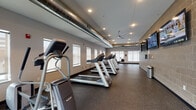 This fitness center is designed to support a comprehensive workout experience across a thoughtfully organized, single-floor layout. The space is divided into distinct exercise zones, including a dedicated cardio area lined with multiple treadmills and additional aerobic machines positioned along a row of windows that allow natural light to fill the room. Exposed industrial-style ductwork runs along the ceiling, complemented by recessed lighting and ceiling fans that contribute to a functional, modern aesthetic. The flooring throughout is durable rubber gym matting, ideal for high-traffic exercise use. A separate strength training area features free weights, adjustable benches, and a multi-station cable machine, with large wall-mounted mirrors that visually expand the space and assist with form during workouts. Wall-mounted flat-screen televisions are positioned throughout the facility for entertainment during exercise. The perimeter walls showcase exposed concrete block construction, lending an industrial character to the space. Natural light enters through multiple windows along the exterior walls, offering views of the surrounding area. The facility is accessed through a solid wood-and-glass door set within the concrete block walls, providing a clear transition into the workout space. Overall, the fitness center offers a well-lit, organized, and fully equipped environment suitable for a wide range of fitness activities.