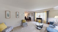 This 1-bedroom, 2-bathroom residence is thoughtfully laid out across a single floor, making efficient use of its space. The combined living and dining area serves as the heart of the home, featuring neutral-toned carpet, soft natural light from large windows dressed with layered curtains, and smooth ceilings. The living area provides ample room for seating arrangements and decorative accents, while the adjacent dining space comfortably accommodates a full dining table with seating for four. A dedicated kitchen features warm wood cabinetry, white appliances including a refrigerator and electric range, light-toned countertops, and vinyl tile flooring — offering a practical and efficient cooking environment. The generously sized primary bedroom is carpeted and benefits from natural light via a window with blinds. It includes access to closet space and connects to a bathroom, creating a comfortable private retreat with warm, neutral wall tones. A second full bathroom features a tiled tub-shower combination. Throughout the residence, the consistent use of neutral walls, soft carpet, and classic finishes creates a cohesive and calm ambiance suited for comfortable everyday living.
