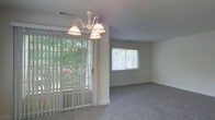 This single-story space features an open living and dining area finished in neutral tones, creating a clean and versatile backdrop. The dining area is anchored by a white chandelier with multiple pendant lights, lending a classic decorative touch to the space. Vertical blinds frame a sliding glass door that opens to an outdoor area, where mature trees are visible, allowing for ample natural light to filter in throughout the day. Adjacent to the dining space, the living area is carpeted in a soft, neutral tone that extends throughout, adding warmth and comfort underfoot. A standard window fitted with blinds provides additional natural light to the living area while maintaining privacy. The walls are painted in a light, muted hue that complements the overall brightness of the space. Ceiling vents indicate a central HVAC system for year-round climate control. The open layout between the two areas promotes a seamless flow, making the space functional for both everyday living and entertaining. Overall, the space presents a simple, well-lit interior with practical features and a connection to the outdoors.