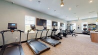 Located at 7800 District Crossing Dr. in Sunbury, Ohio, this fitness center offers a comprehensive exercise facility within a single, well-organized floor. The space is finished with light, neutral-toned walls and patterned carpeting, creating a clean and welcoming environment for residents.

The cardio zone features a row of True-brand treadmills and elliptical machines, each equipped with individual display screens for an interactive workout experience. Wall-mounted flat-screen TVs are positioned throughout, providing entertainment during exercise sessions. Natural light flows in generously through multiple windows fitted with horizontal blinds, keeping the space bright and energizing.

The strength training area is equally well-appointed, featuring a multi-function cable machine, an adjustable weight bench, and a rack of dumbbells spanning a wide range of weights. A dedicated functional training corner houses stability balls of various sizes alongside a TAG-branded rack stocked with medicine balls and kettlebells, catering to a variety of training styles.

Recessed lighting and a decorative ceiling fan complement the overhead illumination, ensuring the space remains well-lit at all times. A water cooler station near the entrance adds a practical convenience. The room's efficient layout maximizes usability while maintaining clear pathways throughout, making this fitness center a versatile and fully functional amenity.