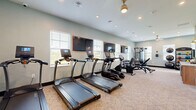 Located at 7800 District Crossing Dr. in Sunbury, Ohio, this fitness center offers a comprehensive exercise facility within a single, well-organized floor. The space is finished with light, neutral-toned walls and patterned carpeting, creating a clean and welcoming environment for residents.

The cardio zone features a row of True-brand treadmills and elliptical machines, each equipped with individual display screens for an interactive workout experience. Wall-mounted flat-screen TVs are positioned throughout, providing entertainment during exercise sessions. Natural light flows in generously through multiple windows fitted with horizontal blinds, keeping the space bright and energizing.

The strength training area is equally well-appointed, featuring a multi-function cable machine, an adjustable weight bench, and a rack of dumbbells spanning a wide range of weights. A dedicated functional training corner houses stability balls of various sizes alongside a TAG-branded rack stocked with medicine balls and kettlebells, catering to a variety of training styles.

Recessed lighting and a decorative ceiling fan complement the overhead illumination, ensuring the space remains well-lit at all times. A water cooler station near the entrance adds a practical convenience. The room's efficient layout maximizes usability while maintaining clear pathways throughout, making this fitness center a versatile and fully functional amenity.