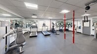 Located at 109 Club Dr in Morganton, North Carolina, this fitness center offers a generously sized, single-floor open layout designed to accommodate a comprehensive range of workout activities. The space is thoughtfully organized to maximize usability, with distinct zones for cardio training, strength machines, and free weights.

The cardio section is well-stocked with multiple treadmills, elliptical trainers, and a stationary bike, all positioned near large windows that allow natural light to filter in and provide views of the surrounding outdoors. The strength training area features a multi-station cable machine system along with adjustable benches, supporting a wide variety of resistance exercises. A dedicated free weight zone includes a full rack of dumbbells in graduated sizes, ideal for both beginners and experienced athletes.

Floor-to-ceiling mirrors line the walls throughout the space, visually expanding the room while allowing users to monitor their form during workouts. The durable rubber flooring provides traction and cushioning underfoot, suited for high-impact activities. Overhead, a drop tile ceiling with recessed fluorescent lighting ensures the entire area is evenly and brightly illuminated.

Distinctive red structural columns punctuate the open floor plan, adding a bold visual accent to the otherwise clean, white-and-gray interior. The overall ambiance is functional, bright, and professional, making this facility a well-rounded destination for fitness enthusiasts in the Morganton area.