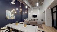 This single-floor space masterfully combines industrial and modern design elements to create a sophisticated, versatile environment. The defining architectural feature is the soaring exposed concrete ceiling, through which visible ductwork and structural components run, lending the space an authentic industrial character.

The interior is divided into distinct functional zones. A dining area anchors one side of the space, centered around a substantial white table paired with sleek dark chairs and illuminated by a striking brass chandelier with multiple candelabra-style bulbs. The adjacent lounge area features plush gray upholstered sofas and armchairs arranged around coffee tables atop a patterned area rug, creating an inviting gathering spot.

A wall-mounted flat-screen television is positioned against a bold navy blue accent wall, which serves as a dramatic focal point and adds depth to the overall design. Light wood flooring runs throughout, providing warmth against the cooler industrial elements above. Numerous pendant lights of varying heights descend from the ceiling, ensuring ample illumination while enhancing the contemporary aesthetic. The result is a well-curated space that balances comfort, style, and functionality.