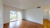 This single-story unit in Miami Gardens, FL offers a well-designed layout with distinct living, dining, kitchen, and bedroom areas. The living room is a standout space, featuring warm wood-style flooring that extends throughout the main areas, a textured ceiling, and a large sliding glass door that opens to views of mature trees and green surroundings, allowing generous natural light to fill the room. The adjoining dining area features a classic chandelier and connects seamlessly to the kitchen via a breakfast bar with granite-style countertops. The kitchen is equipped with dark cabinetry, stainless steel appliances including a range and refrigerator, and tile flooring. A hallway leads to the bedrooms, each carpeted and bright with large windows framing outdoor greenery. The bathroom includes a granite-topped vanity, and a dedicated in-unit laundry closet with a full-size washer and dryer adds everyday convenience. Clean white walls, consistent finishes, and a neutral palette create a cohesive and functional living environment throughout the space.
