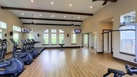 This single-floor fitness center offers a well-appointed workout environment designed for both cardio training and open-floor exercise. The space is defined by soaring ceilings adorned with dark wood decorative beams, which lend a refined architectural character to the room. Recessed lighting and pendant-mounted ceiling fans are distributed throughout, ensuring consistent illumination and comfortable air circulation during workouts.

A row of large, evenly spaced windows along one wall allows abundant natural light to fill the space, providing views of the surrounding greenery and creating an open, inviting atmosphere. The flooring throughout is a warm hardwood-style surface that complements the neutral, light-toned walls.

The cardio zone along one side of the room is equipped with treadmills and other exercise machines, while the expansive open floor area provides ample room for stretching, group fitness, or functional training. A wall-mounted flat-screen television is positioned for easy viewing from multiple areas of the room. Floor-to-ceiling glass-paneled room dividers offer a sleek, modern partition that separates areas while maintaining visual openness. Wall-mounted speakers suggest an integrated audio system to support a motivated workout environment. The overall design balances functionality with a clean, modern aesthetic.