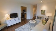 This single-floor residence is designed with an open, connected layout that maximizes the sense of space and flow. The living area serves as the central hub, featuring rich dark hardwood flooring that runs consistently throughout the space, creating visual continuity. A patterned area rug anchors the seating arrangement, which includes a light-toned sofa and accent chair positioned around a round geometric coffee table. A white media console with dark hardware houses the flat-screen television along one wall, complemented by colorful wall art that adds warmth to the neutral palette.

Adjacent to the living area, the kitchen is accessible through an open cutout, revealing light wood cabinetry, dark stone countertops, and stainless steel appliances including a microwave and range. Track lighting on the ceiling provides focused illumination across both spaces. A hallway visible from the living room leads further into the unit, hinting at additional private spaces beyond. The overall ambiance is clean, modern, and functional, with consistent finishes and a cohesive color scheme of whites, neutrals, and warm wood tones throughout.