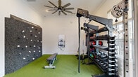 Located at 380 Topwater Dr in Clayton, NC, this single-floor amenity space offers residents a well-rounded lifestyle experience through two distinct areas. The fitness center is comprehensively outfitted with cardio equipment including multiple treadmills and a rowing machine, a multi-functional cable rack, a full dumbbell rack, kettlebells, medicine balls, resistance bands, and a climbing wall. Artificial turf flooring defines the strength-training zone, while neutral carpet covers the cardio area. Ceiling fans, crown molding, large windows overlooking the pool, and a dual water fountain station contribute to a clean, comfortable workout environment. The adjacent covered outdoor lounge area provides a relaxed gathering space with paneled sage-green accent walls, large blue sectional seating, a white round coffee table, and a brick-accented fireplace with a wall-mounted television. Open on one side, the lounge overlooks the community pool and surrounding greenery, blending indoor comfort with outdoor ambiance. A ping-pong table and bench seating add a recreational dimension to the space. Together, these areas form a versatile, resort-style amenity hub for the surrounding residential community.
