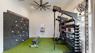 Located at 380 Topwater Dr in Clayton, NC, this single-floor amenity space offers residents a well-rounded lifestyle experience through two distinct areas. The fitness center is comprehensively outfitted with cardio equipment including multiple treadmills and a rowing machine, a multi-functional cable rack, a full dumbbell rack, kettlebells, medicine balls, resistance bands, and a climbing wall. Artificial turf flooring defines the strength-training zone, while neutral carpet covers the cardio area. Ceiling fans, crown molding, large windows overlooking the pool, and a dual water fountain station contribute to a clean, comfortable workout environment. The adjacent covered outdoor lounge area provides a relaxed gathering space with paneled sage-green accent walls, large blue sectional seating, a white round coffee table, and a brick-accented fireplace with a wall-mounted television. Open on one side, the lounge overlooks the community pool and surrounding greenery, blending indoor comfort with outdoor ambiance. A ping-pong table and bench seating add a recreational dimension to the space. Together, these areas form a versatile, resort-style amenity hub for the surrounding residential community.