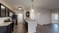 This single-floor residence at Ashton Park offers a thoughtfully designed layout with modern finishes throughout. The kitchen serves as a standout feature, configured in a galley style with rich, dark espresso cabinetry providing ample storage on both sides. Granite countertops run the length of the kitchen, complemented by a tiled backsplash and stainless steel appliances, including a gas range, built-in microwave, refrigerator, and dishwasher. A peninsula with an integrated sink extends into the living area, topped with matching granite and illuminated by pendant lighting, creating a natural division between the kitchen and adjacent dining space.

The open living and dining area is generously sized, featuring dark wood-look flooring that flows consistently through the main living spaces. A ceiling fan and pendant light fixture provide both function and style, while a window admits natural light. The bedroom offers a comfortable retreat with plush carpeting and multiple windows fitted with blinds. The bathroom includes a dark-cabineted vanity with a granite countertop, wood-look flooring, and a full tub and shower combination set within clean, bright surroundings.