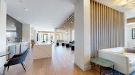This expansive amenity lounge spans a generously sized single floor, offering a thoughtfully designed communal space with distinct functional zones connected by cohesive contemporary styling. The flooring transitions between wide-plank wood-look tile and decorative geometric patterned tile, delineating seating areas while maintaining visual continuity throughout the space.

At the heart of the lounge, vertical wood slat partitions serve as architectural dividers, adding warmth and rhythm without fully enclosing the open-plan layout. A dedicated bar area features a dramatic dark subway tile backsplash, a wall-mounted television, and a substantial white marble-veined countertop lined with leather-and-metal barstools, creating an ideal social hub.

The lounge incorporates multiple seating configurations, including built-in banquette seating along window walls, a relaxed living area with a white sofa, velvet accent chairs, and patterned area rugs, as well as intimate groupings of ottomans and occasional tables. Bold teal wainscoted accent walls contrast elegantly with the predominantly white palette.

Floor-to-ceiling black-framed windows and glass doors flood the interior with natural light and provide views of the surrounding outdoor courtyard. Layered lighting, including globe pendants, flush-mount fixtures, and recessed downlights, ensures a well-lit and inviting ambiance throughout.