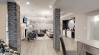 This single-floor space offers a thoughtfully designed open-concept layout that seamlessly blends style and functionality. The main living area is anchored by a striking stacked-stone column that rises from floor to ceiling, serving as a bold architectural focal point. Light hardwood flooring flows throughout, lending warmth and continuity to the space.

The living area is well-appointed with a mix of patterned and neutral seating arranged around a tufted ottoman, complemented by a classic fireplace and a wall-mounted television. A sputnik-style chandelier with a starburst silhouette hangs overhead, adding a modern touch beneath the recessed-lit ceiling. Natural light filters in through white shuttered windows, brightening the neutral gray and white color palette.

Adjacent to the main seating area, a dining space provides room for gatherings, while a built-in kitchenette tucked into a darkly accented alcove offers a sink, countertop appliances, and cabinetry — making the space well-suited for entertaining. Decorative mirrors and contemporary wall art add curated finishing touches throughout. Overall, the space exudes a polished, contemporary ambiance that is both inviting and visually refined.