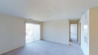 This single-floor space presents a clean, open layout with a bright and airy atmosphere. The main living area is finished with plush wall-to-wall carpeting in a neutral tone, providing a comfortable underfoot feel throughout. A prominent sliding glass door, dressed with vertical blinds, serves as both a natural light source and an access point to the outdoors, offering views of surrounding greenery. The large door allows ample daylight to fill the room, enhancing the sense of openness within the space.

The walls are finished in a soft, warm off-white palette that complements the light carpeting and contributes to the overall bright, neutral aesthetic. A ceiling-mounted HVAC vent ensures climate control throughout the area. The smooth, flat ceiling maintains a clean and uncluttered look above.

A doorway visible from the main living area leads to an additional room, suggesting a connected layout that allows for flexible use of the space. The overall design is simple and functional, with minimal architectural embellishments, making it highly adaptable to a variety of furnishing styles and personal preferences. The combination of natural light, neutral finishes, and a straightforward floor plan gives this space a welcoming and practical character.