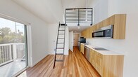 This thoughtfully designed two-story loft in Seattle, WA offers a smart and efficient layout spread across two floors. The main level features an open-plan kitchen and living area bathed in natural light, thanks to large sliding glass doors that lead directly to a private balcony with neighborhood views. The kitchen is outfitted with warm wood-tone cabinetry, white quartz countertops, stainless steel appliances including a refrigerator, dishwasher, built-in microwave, and an electric cooktop — all seamlessly integrated along a single wall for a clean, modern aesthetic. Light hardwood flooring runs throughout, complementing the crisp white walls and contemporary finishes. Soaring double-height ceilings in the living area create a striking sense of openness and volume, amplified further by a skylight that draws in additional natural light. A sleek black metal ladder leads to the upper loft level, which serves as the bedroom and is bordered by a modern horizontal-rail railing, offering an open view down to the main floor. The full bathroom is finished with matching wood-tone cabinetry, a white vanity, and a frosted glass shower enclosure. This residence combines minimalist design with functional living in a compact yet surprisingly airy footprint.