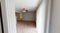 This single-floor residence offers a practical and comfortable layout across several well-defined living spaces. The open living area is a standout feature, highlighted by neutral gray walls, plush wall-to-wall carpeting, and a ceiling fan with a built-in light fixture. Floor-to-ceiling vertical blinds line a large sliding glass door, allowing abundant natural light to fill the space while providing access to the outdoors. The living area flows seamlessly from a secondary space defined by a pass-through breakfast bar with a dark wood upper cabinet and subway tile backsplash, offering a convenient connection to the kitchen. The galley-style kitchen is appointed with rich dark wood cabinetry, black appliances including an electric range and refrigerator, light countertops, and warm hardwood flooring — creating a functional and cohesive cooking environment. The bedrooms continue the neutral, carpeted aesthetic, each equipped with ceiling fans and natural light from windows fitted with blinds. At least one bedroom features access to a walk-in closet, adding valuable storage. Clean white trim and consistent gray wall tones throughout the space contribute to a unified, move-in-ready ambiance suited for everyday living.