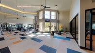 This expansive fitness center is designed to accommodate a wide range of workout styles within a single, thoughtfully organized space. The main gym area impresses with dramatic double-height ceilings and abundant natural light streaming through tall windows and glass doors that open to an outdoor area. Large ceiling fans and recessed lighting complement the bright, airy atmosphere. The flooring features a colorful patchwork carpet tile pattern in neutral, blue, orange, and black tones, adding visual interest throughout the open floor plan.

The main workout area is well-stocked with a full rack of dumbbells, strength machines, cardio equipment including ellipticals and stair climbers, and floor-to-ceiling mirrors with wall-mounted televisions. A dedicated functional training zone offers TRX suspension systems, yoga mats, and a cable machine, while a wooden storage console with cubbies and athletic-themed artwork anchors the entry area.

A separate, glass-enclosed spin studio features wood-look flooring, a row of cycling bikes, medicine balls, and mirrored walls, creating an intimate yet visually open environment. Double glass entry doors connect the fitness center to an outdoor terrace, further enhancing the sense of openness and connectivity throughout the facility.
