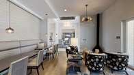 This well-appointed clubhouse is designed to serve as a sophisticated communal gathering space, thoughtfully arranged across a single floor. The interior features warm hardwood-style flooring throughout, crisp white ceilings with coffered detailing, and a neutral gray and white color palette that lends the space a clean, modern aesthetic.

The main lounge and dining area is anchored by a generously sized built-in banquette upholstered in soft gray, paired with coordinating dining chairs at dark wood tables — ideal for casual meals or social gatherings. Overhead, a striking sputnik-style crystal chandelier and complementary pendant lights serve as dramatic focal points, elevating the overall design.

A freestanding black conical fireplace adds a bold architectural element to the lounge zone, where sculptural lattice-patterned armchairs and a low cocktail table create an inviting conversation area. Large floor-to-ceiling glass doors flood this corner with natural light and offer open views of the surrounding outdoor community spaces.

Toward the rear, a stylish bar area features a granite-topped counter with tufted high-back stools, a patterned accent wall, and a wall-mounted television — perfect for entertaining. The seamless flow between zones makes this clubhouse a versatile and visually engaging amenity space.