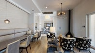 This well-appointed clubhouse is designed to serve as a sophisticated communal gathering space, thoughtfully arranged across a single floor. The interior features warm hardwood-style flooring throughout, crisp white ceilings with coffered detailing, and a neutral gray and white color palette that lends the space a clean, modern aesthetic.

The main lounge and dining area is anchored by a generously sized built-in banquette upholstered in soft gray, paired with coordinating dining chairs at dark wood tables — ideal for casual meals or social gatherings. Overhead, a striking sputnik-style crystal chandelier and complementary pendant lights serve as dramatic focal points, elevating the overall design.

A freestanding black conical fireplace adds a bold architectural element to the lounge zone, where sculptural lattice-patterned armchairs and a low cocktail table create an inviting conversation area. Large floor-to-ceiling glass doors flood this corner with natural light and offer open views of the surrounding outdoor community spaces.

Toward the rear, a stylish bar area features a granite-topped counter with tufted high-back stools, a patterned accent wall, and a wall-mounted television — perfect for entertaining. The seamless flow between zones makes this clubhouse a versatile and visually engaging amenity space.