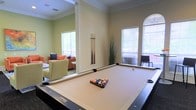 This single-floor amenity space is thoughtfully designed to offer both recreation and relaxation in a bright, well-appointed setting. The centerpiece of the space is a full-sized billiards table, positioned in a dedicated game room with elevated ceilings accented by elegant crown molding. A wall-mounted cue rack keeps equipment neatly organized, while multiple windows with horizontal blinds allow generous natural light to fill the room. An arched window adds a classic architectural touch, enhancing the refined character of the space. Adjacent to the billiards area, an open lounge offers a comfortable seating arrangement featuring plush armchairs and a vibrant coral sofa, creating a warm and inviting atmosphere for socializing. The lounge is carpeted and styled with decorative accents, providing a relaxed contrast to the game room's harder surfaces. Neutral, light green walls throughout the space contribute to an airy and welcoming ambiance. The open layout between the game room and lounge encourages a seamless flow, making this space well-suited for both casual gatherings and recreational activities.