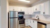 Located at 1747 Alexander Springs Ln in Wake Forest, NC, this single-floor unit offers a well-designed layout with modern finishes and a cohesive, neutral color palette. The kitchen is a standout feature, outfitted with crisp white shaker-style cabinetry accented by matte black hardware, a classic white subway tile backsplash, stone-look countertops, and a full suite of stainless steel appliances including a refrigerator, electric range, over-the-range microwave, and dishwasher. A double-basin sink with a matte black faucet completes the space. Wood-look luxury flooring extends throughout the unit, providing a warm, cohesive feel that ties the rooms together. The open living area is finished with crown molding, neutral gray walls, and abundant natural light from large windows, creating an airy and inviting atmosphere. The bedroom features a ceiling fan with lighting and a window that brings in additional natural light. The bathroom is appointed with wood-look tile flooring, a tub-shower combination with brushed gold fixtures, and a built-in linen closet for added storage. A dedicated laundry room with wire shelving accommodates a full-size side-by-side washer and dryer, adding everyday convenience to this thoughtfully finished single-level home.