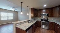 This single-floor residence offers a thoughtfully designed open-concept layout that seamlessly connects the kitchen and living areas. The kitchen is a standout feature, showcasing rich dark wood cabinetry, granite countertops, and a full suite of stainless steel appliances including a refrigerator, range, and over-the-range microwave. A peninsula with pendant lighting overhead provides additional prep space and serves as a natural divider between the kitchen and the adjoining living area. The living space is bright and welcoming, featuring warm hardwood flooring, neutral-toned walls, and a ceiling fan that enhances air circulation. Natural light flows in through large windows fitted with blinds, offering views of the surrounding neighborhood. The bedrooms continue the hardwood flooring and neutral palette, each equipped with ceiling fans and windows that bring in abundant daylight. The bathroom is well-appointed with a granite-topped dark wood vanity, large mirror, and light tile flooring. Notably, the bathroom includes accessibility features such as grab bars near the toilet, making the space inclusive and functional. Throughout, the consistent use of hardwood floors, granite surfaces, and dark wood cabinetry creates a cohesive and polished aesthetic.