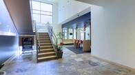 Located at 415-425 N Front St in Columbus, Ohio, this commercial space makes a strong first impression with its expansive, light-filled lobby. The entrance area features striking stone tile flooring in a varied pattern, lending a sense of permanence and sophistication to the space. Soaring ceilings with tall windows flood the area with natural light, creating an open and welcoming atmosphere.

A handsome wood-and-glass staircase anchors the lobby, offering both functional access and an architectural focal point. Pendant lighting descends from the high ceiling, adding warmth and a contemporary touch. Potted greenery is thoughtfully placed throughout, softening the clean lines of the interior.

Adjacent to the main lobby is a collaborative area defined by a richly colored, dark blue accent wall and carpeted flooring. This zone is furnished with a long wood-toned counter and coral-hued bar seating, creating an inviting space well-suited for informal meetings or breakout sessions. Warm pendant lights and framed mirror panels enhance the area's modern yet approachable aesthetic.

Overall, the space blends industrial and contemporary design elements, offering a professional and dynamic environment ideal for business or office use in the vibrant downtown Columbus corridor.