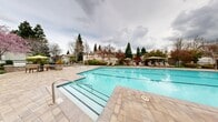Located at 1300 Burton Dr in Vacaville, California, this two-story residential community features a generously sized outdoor pool and spa area that serves as a central gathering space for residents. The large, rectangular lap pool is defined by clean lane markings and is framed by an expansive paver deck finished in neutral tones, providing ample room for lounging and circulation. Entry to the pool is made accessible via wide, gradual steps with sturdy handrails on both ends of the pool.

Adjacent to the main pool sits a separate spa, offering a more intimate relaxation option. The surrounding deck is outfitted with rows of cushioned lounge chairs and shaded umbrella tables, creating comfortable spaces for outdoor leisure. Mature trees — including towering pines and seasonal flowering trees — line the perimeter, providing natural shade and a lush, landscaped backdrop that enhances the overall ambiance.

A well-maintained clubhouse building borders one side of the pool area, featuring arched windows, stucco exterior, and a gated entry point. Manicured shrubs and colorful plantings are integrated throughout the space, adding vibrancy and a sense of privacy. The pool area is fully enclosed by a wrought iron-style fence, ensuring security while maintaining an open, airy feel. The overall setting is clean, inviting, and thoughtfully designed for outdoor enjoyment.