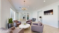 Located at 10105 Wornall Rd in Kansas City, MO, this single-floor space presents a clean, contemporary design characterized by soft neutral tones, light wood-look tile flooring, and crisp white crown molding throughout. The open layout flows seamlessly between distinct functional zones, creating an inviting and highly versatile environment.

The lounge area serves as a central gathering space, furnished with plush upholstered armchairs arranged around a sleek white round coffee table. A modern geometric chandelier anchors the room overhead, complemented by recessed lighting. Large windows fitted with horizontal blinds flood the space with natural light and offer views of the surrounding greenery outdoors.

Adjacent to the lounge, a dining area features a white rectangular table with contemporary chrome-legged chairs. A striking stacked stone accent wall provides a bold textural contrast to the otherwise minimalist palette, with a built-in niche displaying greenery. An identical chandelier ties the two areas together cohesively.

Built-in workstations with white desks and wall-mounted monitors are thoughtfully integrated into alcoves along the walls. Glass-paneled partition walls and French doors define additional rooms beyond, maintaining an open, airy feel while offering functional separation. The overall ambiance is polished, light-filled, and professionally appointed.