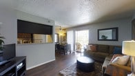 This single-floor residence offers a thoughtfully designed open-concept layout that seamlessly connects the living, dining, and kitchen areas. The living room is a welcoming space, highlighted by rich hardwood-style flooring, a plush area rug, and a ceiling fan that adds both style and function. A large sliding glass door floods the room with natural light and opens to a private balcony, offering pleasant outdoor views of surrounding greenery. The living area flows naturally into the dining space, which comfortably accommodates a full dining set beneath a ceiling fan with a warm light fixture. A pass-through window connects the dining area to the kitchen, creating a convenient and social layout ideal for entertaining. The kitchen features warm countertops and is visible through the pass-through, giving the overall space an interconnected feel. Textured ceilings add a classic architectural touch throughout, while neutral grey walls provide a versatile backdrop that complements a variety of interior styles. The combination of natural light, open sightlines, and cohesive flooring gives this residence a comfortable, functional, and inviting ambiance suited for everyday living.