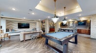Located at 7490 Dwell Well Way in Winter Park, Florida, this well-appointed community clubroom offers a generously sized, open-plan gathering space designed for both recreation and relaxation. The interior is defined by rich wood-look tile flooring, textured stone-clad columns, and a tray ceiling with recessed lighting that adds architectural depth and warmth throughout.

The game area serves as a central focal point, featuring a full-size billiards table and a shuffleboard table, both illuminated by stylish industrial-style pendant lights. Large floor-to-ceiling windows along multiple walls flood the space with natural light and provide open views of the surrounding landscaped grounds.

A built-in lounge nook offers a cozy retreat, complete with a large wall-mounted flat-screen TV set against a bold chevron-patterned accent wall and a cushioned bench seat accented with decorative pillows. Adjacent seating areas provide comfortable spaces for socializing.

The wet bar area is a standout feature, showcasing a striking granite-topped island with upholstered bar stools, globe pendant lighting, stainless steel under-counter refrigerators, a draft beer tap, and wall-mounted televisions — creating a sophisticated entertaining hub within the space.