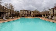 This residential community showcases a blend of traditional architectural design and thoughtfully curated outdoor amenities. The exterior buildings feature classic red brick facades accented with neutral stucco detailing, multi-pane windows, and private balconies that add depth and character to the overall aesthetic.

At the heart of the community lies a generously sized swimming pool with striking turquoise water, complemented by a built-in spa or wading feature at its center. The pool deck is finished with warm-toned brick pavers and outfitted with lounge chairs, providing ample space for relaxation. A covered cabana with white columns and a pitched roof offers shaded seating and serves as an attractive focal point along the poolside.

Beyond the main pool area, a secondary outdoor courtyard features additional brick paver hardscaping with umbrella-shaded tables and chairs, ideal for casual gatherings. Mature trees and carefully manicured landscaping, including trimmed hedges, colorful plantings, and well-maintained lawn areas, frame the buildings and pathways throughout the property, creating a welcoming, resort-like atmosphere.

The community entrance is marked by an elegant stucco-clad structure with decorative wrought iron gating, large multi-pane windows, and ornamental container plantings, establishing a refined first impression that reflects the overall character of the property.