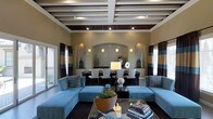This single-floor space showcases a thoughtfully designed open-plan layout that seamlessly connects the living and dining areas into one cohesive, elegant environment. The living area is anchored by a pair of plush, sky-blue sectional sofas arranged around a sleek glass coffee table, creating a welcoming and stylish seating arrangement. A rich brown and blue striped drapery frames the windows on either side, allowing natural light to filter in while complementing the room's sophisticated color palette. The ceiling is a standout architectural feature, boasting a coffered design with dark wood beams set against a crisp white grid, adding depth and visual interest to the space. Recessed lighting is integrated throughout, ensuring the interior is warmly and evenly illuminated. The dining area, visible through a graceful arched alcove, features a dark wood table surrounded by upholstered chairs, with pendant lights suspended above to create an intimate atmosphere. Decorative niches with accent lighting flank the archway, adding an elegant, gallery-like quality to the transition between spaces. The overall ambiance is refined and inviting, blending contemporary design elements with warm, neutral tones and bold accents for a balanced and polished aesthetic.