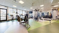 This fitness center offers a thoughtfully designed single-floor layout that accommodates a comprehensive range of exercise equipment and training zones. The space features a distinctive multi-tone flooring pattern in green, yellow, and neutral tones, clearly delineating different workout areas while adding visual interest throughout.

Along the perimeter, floor-to-ceiling windows flood the cardio zone with natural light, where a row of modern treadmills and elliptical machines are positioned to provide outdoor views during workouts. A dedicated functional training area includes TRX suspension systems, battle ropes, stability balls, a BOSU trainer, and a flat bench, complemented by vivid nature-themed murals depicting rock climbing and forested landscapes.

The strength training section houses a multi-station cable machine, a full dumbbell rack with mirrored walls, and various resistance machines suited for both upper and lower body exercises. A water fountain and open floor space near the entrance add practical convenience. The drop ceiling with recessed panel lighting ensures consistent, even illumination throughout, while bold navy accent walls and branded flooring elements contribute to a cohesive, energizing atmosphere.