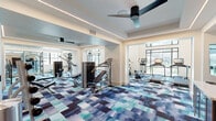 Located at 13220 North Scottsdale Road in Scottsdale, Arizona, this well-appointed fitness center offers a generously sized, single-floor exercise space designed to accommodate a full range of workout needs. The room makes a strong first impression with soaring double-height ceilings in key areas, floor-to-ceiling windows that flood the interior with natural light, and striking views of the outdoors. A bold, multi-toned blue and teal patterned carpet adds a vibrant, contemporary energy underfoot throughout the entire space.

The fitness center is comprehensively equipped for both cardio and strength training. A dedicated cardio zone features Life Fitness ellipticals, treadmills, and a stair climber, all positioned to take advantage of the abundant natural light and outdoor views. The strength training area includes a cable machine system, multi-use weight benches, and a full rack of dumbbells, catering to a wide variety of exercise routines.

Architectural details elevate the space beyond a standard gym. Recessed linear LED lighting trims the tray ceilings in the lower sections, while a geometric chandelier and modern ceiling fans provide visual interest in the taller areas. Large wall mirrors expand the sense of space and aid in proper form. A vibrant landscape artwork adds a decorative focal point, completing this polished and functional fitness environment.