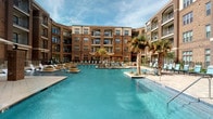 This outdoor amenity space centers around a generously sized resort-style swimming pool that serves as the focal point of an enclosed courtyard. The pool features crystal-clear turquoise water and is bordered by a wide, paver-tiled deck that provides ample space for lounging. Scattered throughout the deck are comfortable lounge chairs and seating areas shaded by large umbrellas, creating an inviting atmosphere for relaxation. Mature palm trees and drought-tolerant plantings are thoughtfully integrated throughout the space, adding a tropical aesthetic to the environment.

The courtyard is framed on multiple sides by a curved, multi-story brick building with arched windows, private balconies, and ground-level access points, creating a sense of enclosure and privacy. A pergola-style structure near the pool's edge provides an additional shaded gathering area, while raised planters with succulents and ornamental grasses add landscaping detail at water level.

Supporting the outdoor space is a clean, well-maintained restroom facility finished with classic white subway tile in a herringbone pattern, sage-green upper walls, framed mirrors, and stainless steel fixtures. The restroom is ADA-accessible, equipped with grab bars and a wall-mounted baby changing station, ensuring convenience and inclusivity for all users.